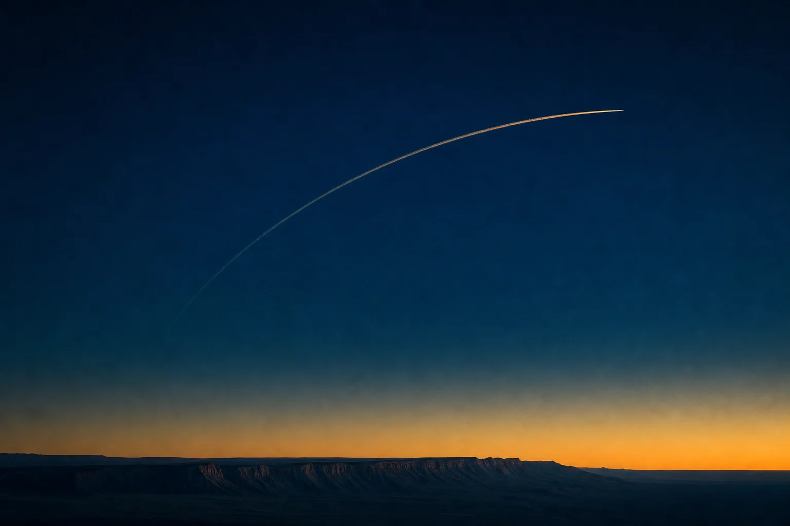Single aircraft contrail arcing across a deep twilight sky above a desert escarpment